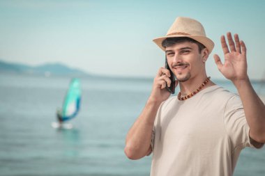 Sahilde cep telefonuyla el sallayan genç bir adam.