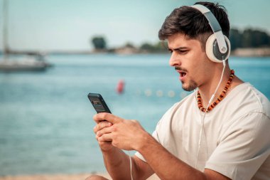 Sahilde cep telefonu kullanan, kulaklıklı, şaşırmış bir genç adam.