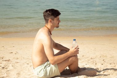 Genç adam elinde bir şişe suyla sahilde denize bakıyor.