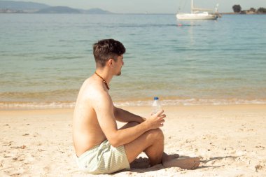 Genç adam elinde bir şişe suyla sahilde denize bakıyor.