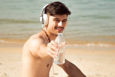 Plajda rahat bir adam kulaklıkla müzik dinliyor ve bir şişe su içiyor.