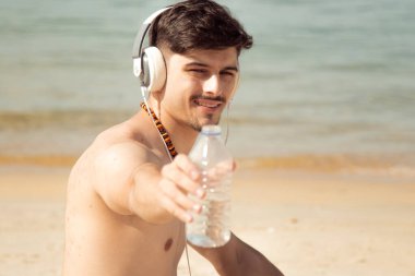 Plajda rahat bir adam kulaklıkla müzik dinliyor ve bir şişe su içiyor.