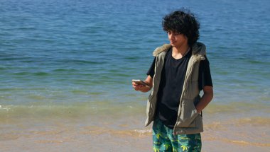 Genç adam sahilde yürüyor ve cep telefonunu kullanıyor.