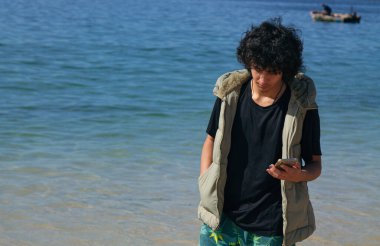 Genç adam sahilde yürüyor ve cep telefonunu kullanıyor.