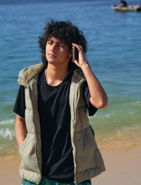 Latin kökenli genç bir adam sahilde yürüyor ve telefonla konuşuyor.