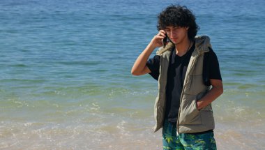 Sahilde cep telefonuyla gülümseyen genç adam.