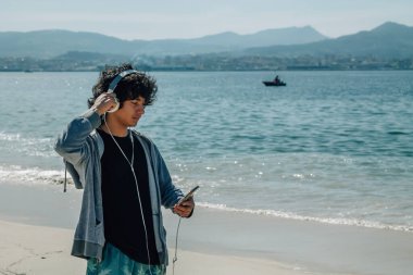 Kulaklıklı ve telefonlu genç adam plajda rahatça yürüyor.