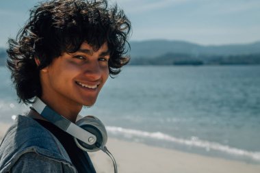 Kulaklıklı genç adam denizin önünde gülümsüyor.