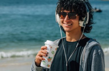 Kulaklıklı genç adam plajda dinleniyor ve bir şeyler içiyor.