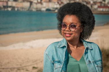 Kıvırcık saçlı, güneş gözlüğü takan ve ceketli bir kadın kumsalda gülümsüyor..
