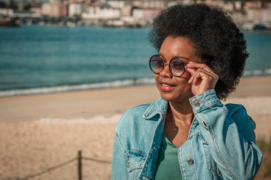 Kıvırcık saçlı, güneş gözlüğü takan ve ceketli bir kadın kumsalda gülümsüyor..