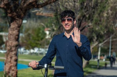 Elektrikli scooterlı genç adam sokakta yürüyor ve el sallıyor.