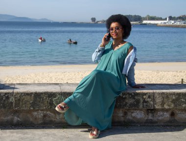 Akıllı telefonlu bir kadın kumsalda oturmuş konuşuyor.