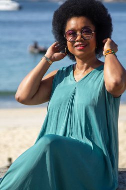 Güneş gözlüklü afro saçlı kadının portresi denizin önünde gülümsüyor.