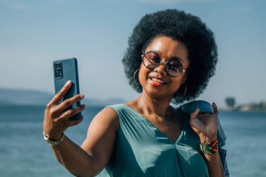 Genç bir kadın özçekim yapıyor ya da sahilin arka planında akıllı telefonlu canlı bir video çekiyor.