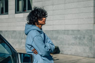 Sokakta bir arabaya yaslanmış kollarını kavuşturmuş genç bir adam.