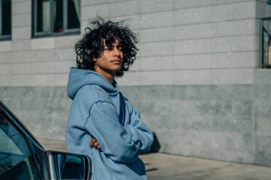 Sokakta bir arabaya yaslanmış kollarını kavuşturmuş genç bir adam.