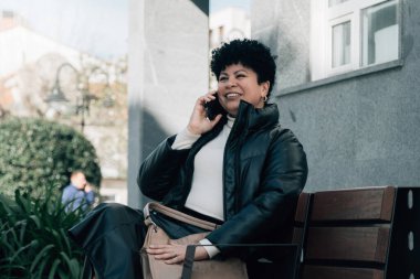 Orta yaşlı bir kadın bankta oturuyor ve cep telefonuyla konuşuyor.