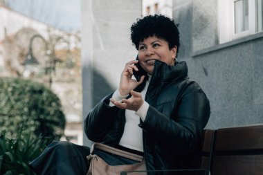 Orta yaşlı bir kadın bankta oturuyor ve cep telefonuyla konuşuyor.