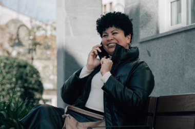 Orta yaşlı bir kadın bankta oturuyor ve cep telefonuyla konuşuyor.