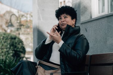 Orta yaşlı bir kadın bankta oturuyor ve cep telefonuyla konuşuyor.