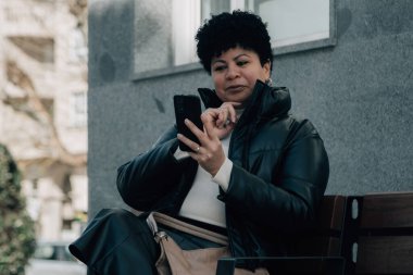 Orta yaşlı bir kadın cep telefonunu kullanarak bankta oturuyor.