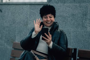 Orta yaşlı bir kadın cep telefonunu kullanarak bankta oturuyor.