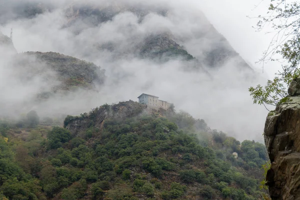 A house in the mountains on a hill surrounded by green forests. The house and the mountain are shrouded in mist and clouds.