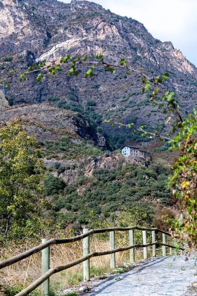 A hiking trail in Andorra, which is paved and protected by a wooden railing. In the background you can see mountains and a house in the mountain. You can also see many plants.