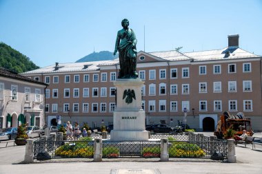 Salzburg, Salzburg - Avusturya - 06-17-2021: Çiçekler ve bitkilerle çevrili bir meydanda Mozart heykeli. Etiketli bir kaide üzerinde figür.