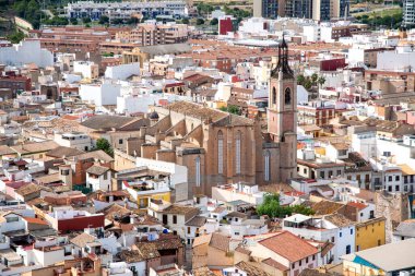 Sagunt, Valencia - İspanya - 09-07-2021: Merkezinde büyük bir kilise, arkasında dağlar ve mavi gökyüzü olan Sagunt üzerinde bir manzara. Birçok çatıda parmaklıklar var..