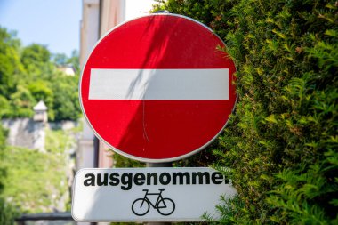 Ortasında beyaz çubuk olan kırmızı yuvarlak bir tabela. Bu da yola doğru sürmene izin verilmediği anlamına geliyor. Tabelanın altında bisikletlerin dışlandığına dair bir işaret var. İşaretler yeşil bir çalılığın yanında.