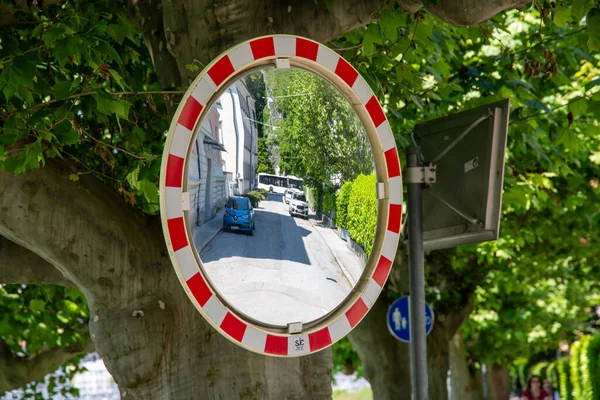 A round traffic mirror with a red and white border and a street in the mirror image where cars are parked. The mirror image is distorted and is fixed under a tree....