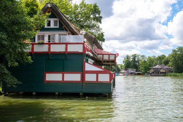Wusterhausen, Brandenburg - Almanya - 06-28-2022: Değişik daireleri ve balkonları olan, gölün huzurlu manzarasına sahip, yeşil suları olan bir göldeki tahta ayaklıklar üzerindeki qaint kayıkhanesi