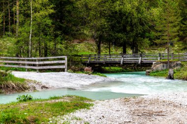 Alpen, Güney Tyrol - İtalya - 06-07-2021: Ahşap köprü Alp Ormanı 'ndaki taşlı bankaların bulunduğu geniş, berrak turkuaz bir dere üzerinden geçiyor