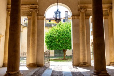 Vicenza, Venetien - İtalya - 06-12-2021: Sessiz, yemyeşil bir ağacı olan güneşli bir avlu, sütunları olan klasik bir portico tarafından çerçevelenmiş ve süslü döşenmiş bir lamba ve açık kapılı çit.
