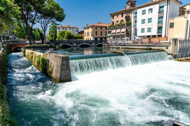 Treviso, Venetien - İtalya - 06-13-2021: Arka planda tarihi binalar bulunan Treviso 'da akarsu ve balık merdiveni bulunan bir Weir şelalesinin canlı görüntüsü