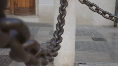 Beton sütunlar arasında asılı, kalın, pas renginde demir bir zincir üzerine bakın. Tüm sahne kentsel alanda geçiyor.