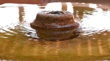 Su yüzeyinde pas renginde kabarcıklar oluşturan yuvarlak bir çeşme. Işık suya yansıyor ve buradan geçen insanları görebilirsiniz.