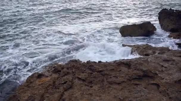 岩の沿岸とその周辺に沿ってその方法を彫刻する岩の海岸線に衝突する波 海はわずかに教会化され 水は白く泡立っている — ストック動画