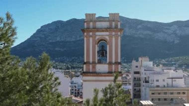 Denia, Alicante - İspanya 11-04-2023: Denia 'da üzerinde kilise çanları olan beyaz bir kilise kulesi. Arka planda mavi bir gökyüzü ve yüksek bir dağ var.