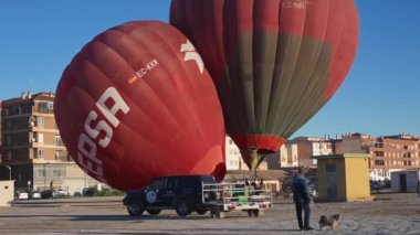 Guadix, Granada - İspanya 01-25-2024: İki kırmızı sıcak hava balonu, biri yükselip diğerini bir kenara itiyor, bir şehir manzarasına karşı. Ön planda, balonların taşıyıcısı..