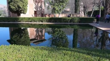 Granada, Granada - İspanya - 01-27-2024: Alhambra bahçesindeki ayna pürüzsüz bir su havzasının üzerinde düzgünce kırpılmış çalılarla kaplı bir tava. Arka planda, ziyaretçiler patika boyunca yürür.