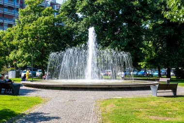 Arona, Piemont - İtalya - 07-04-2024: Arona 'da yeşil ağaçlar ve oturma alanlarıyla çevrili yüksek su jeti bulunan bir halk parkı çeşmesi.
