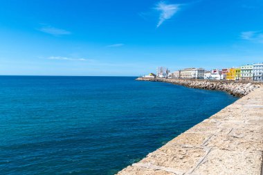 Cadiz, Cadiz - İspanya - 05-06-2024: Evleri ve mavi denizi olan Cadiz 'in kıyı manzarası, berrak gökyüzü ve sakin sular altında bir taş gezinti alanı boyunca.