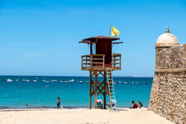 Cadiz, Cadiz - İspanya - 05-06-2024: İspanya 'nın Cadiz kentindeki plajda bulunan cankurtaran kulesi, sarı bir bayrak ve mavi denizin arka planında bulunan botlarla.