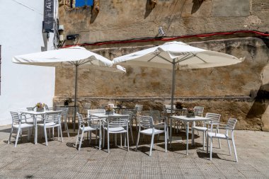 Cordoba, Cordoba - Spain - 04-08-2024: An outdoor cafe setup with tables and umbrellas next to a weathered wall in Cordoba. clipart