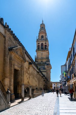 Cordoba, Cordoba - Spanien - 04-08-2024: Mezquita-Katedral Kulesi dar caddenin üzerinde yükseliyor, insanlar kaldırım taşlarından geçerken gün ışığıyla aydınlatılıyor.