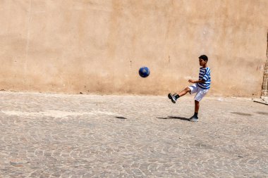 Cordoba, Cordoba - İspanya - 04-08-2024: Kumlu bir duvarın önünde boş bir asfalt alanda futbol oynayan bir çocuk.