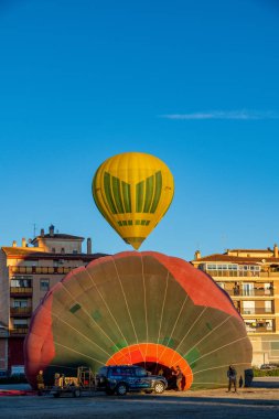 Guadix, Granada - İspanya - 01-24-2024: Sarı bir sıcak hava balonu, yerde kırmızı ve yeşil segmentlerle dolu kısmen dolu büyük bir balonun üzerinde yüzüyor.